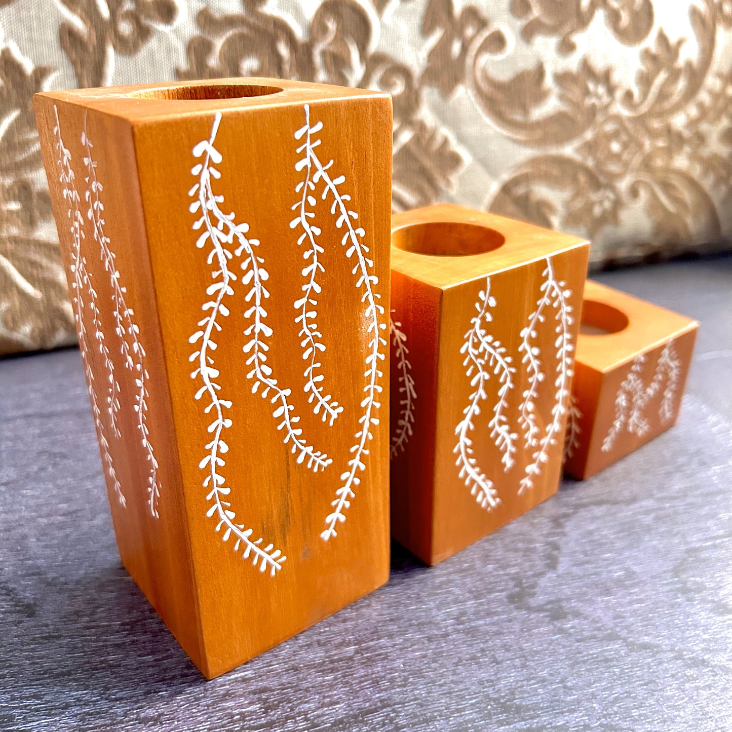 Handcrafted set of 3 candle holders made of Natural solid mahogany wood is a perfect way to make your rooms cozy and beautiful. Hand-drawn with beautiful motifs when placed on a coffee/dining/corner table, accents the beauty,  brightens the space making the room welcoming, calm and peaceful. These candle holders of different heights could be arranged in any way as per the space it is kept. 